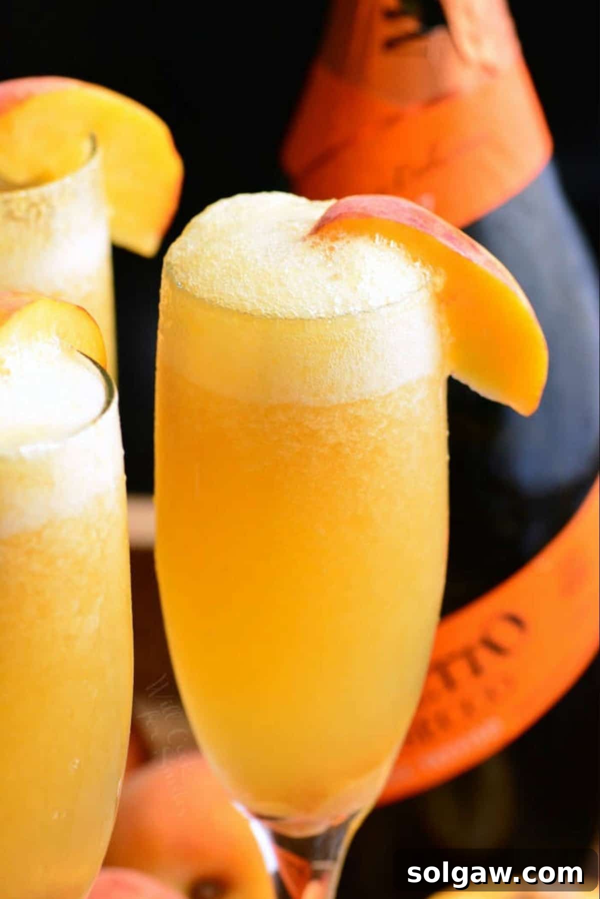 A champagne flute filled with a vibrant orange peach Bellini cocktail, with more glasses blurred in the background, signaling a celebratory mood.