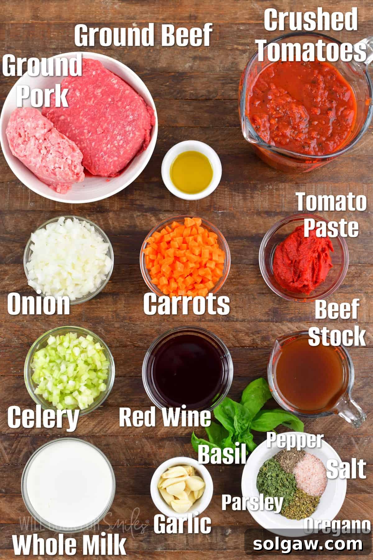 A selection of fresh ingredients laid out on a wooden board for making Bolognese sauce, including onions, carrots, celery, ground meats, garlic, canned tomatoes, and fresh herbs.