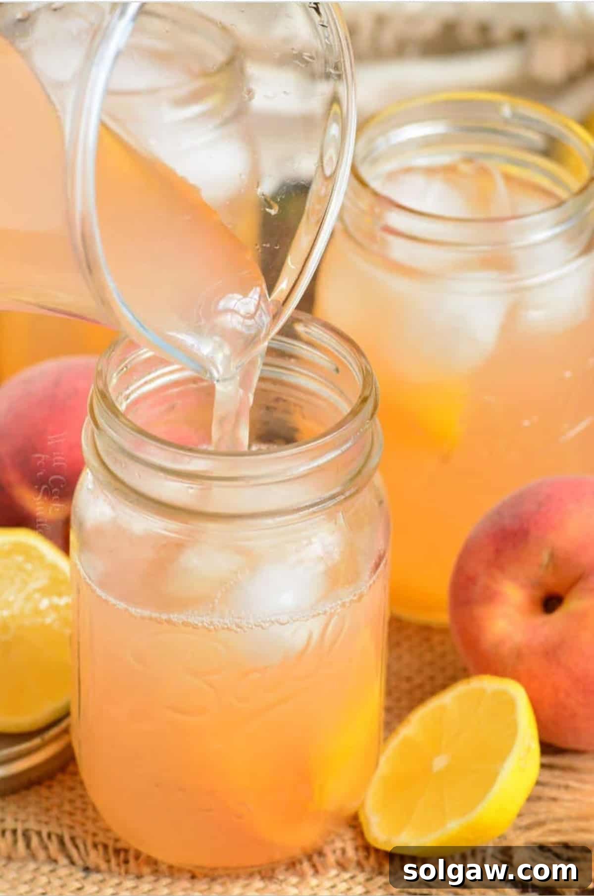 A glass pitcher filled with golden peach lemonade, being poured into a mason jar with ice, promising cool refreshment on a hot day.