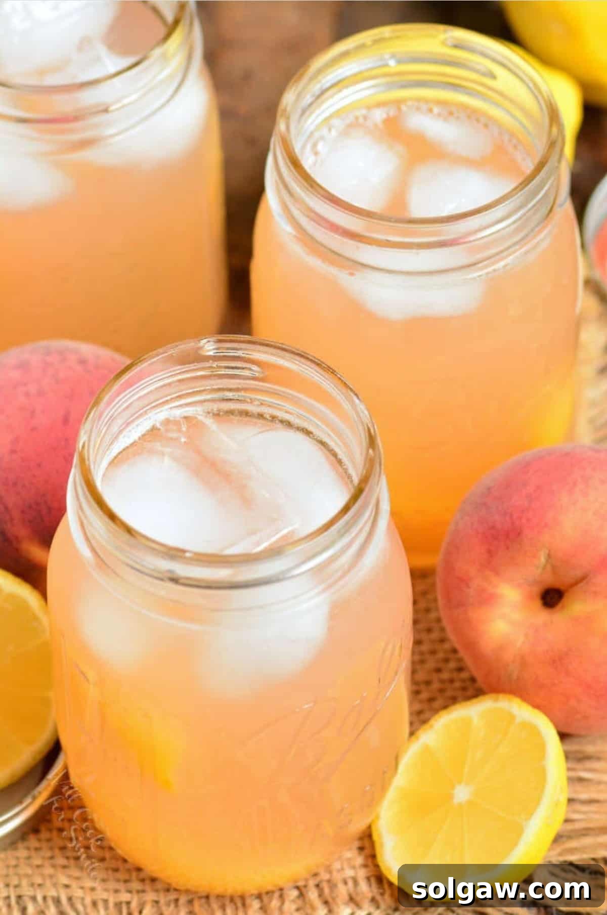 Three inviting mason jars filled with glistening peach lemonade and ice, adorned with fresh peach slices and a mint sprig, awaiting a refreshing indulgence.
