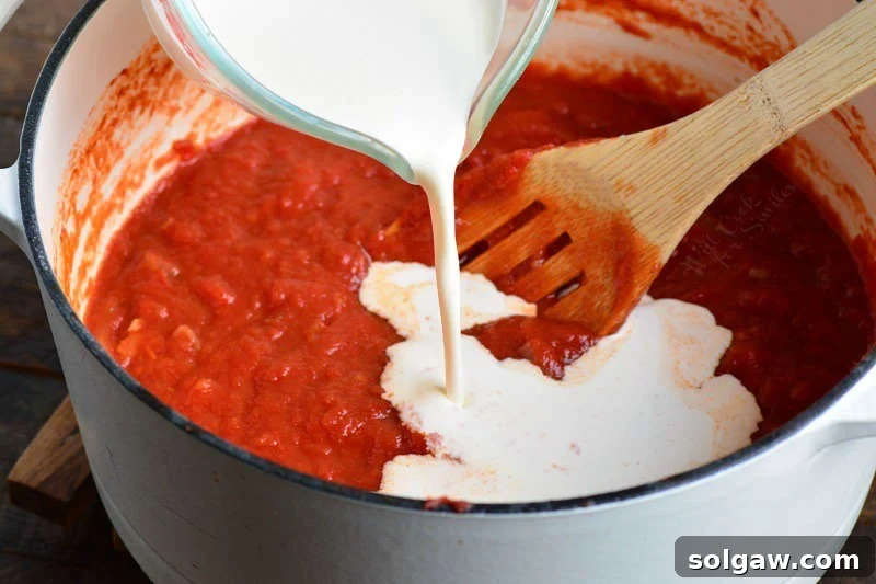 A chef gently pouring rich heavy cream into a simmering pot of vibrant red vodka sauce in a stock pot