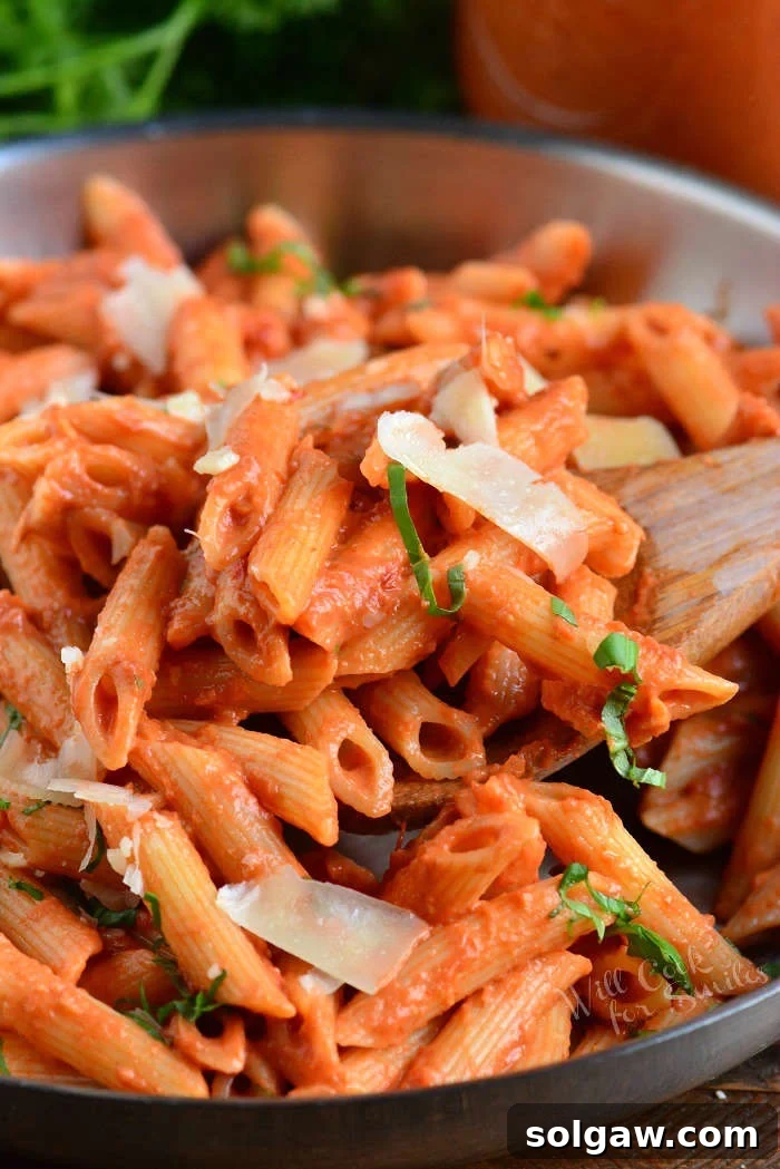 Close-up of creamy vodka sauce coating penne pasta in a pan, with a wooden spoon for serving, garnished with fresh basil