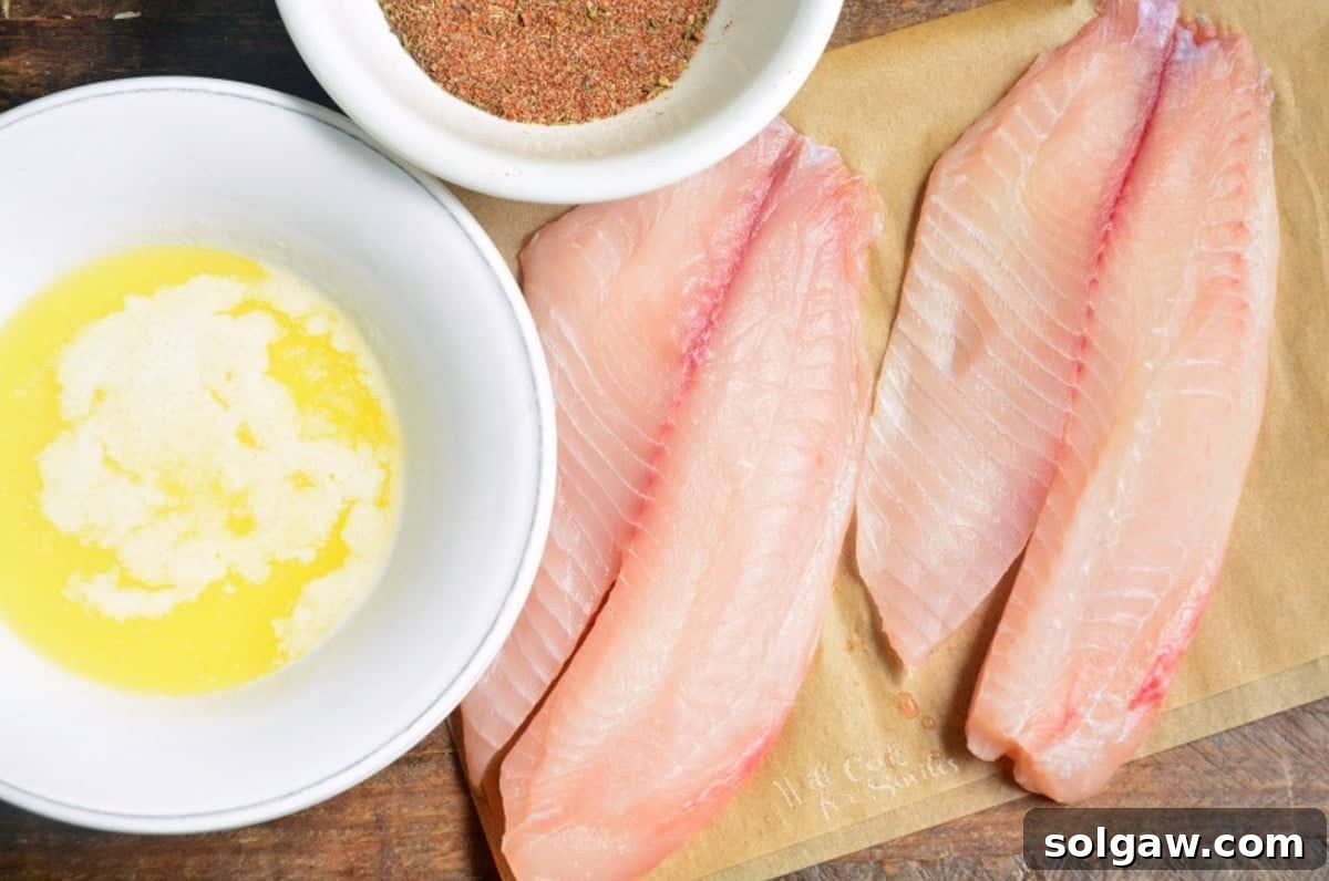 Two pieces of tilapia on parchment paper with a bowl of melted butter and blackening seasoning next to it.