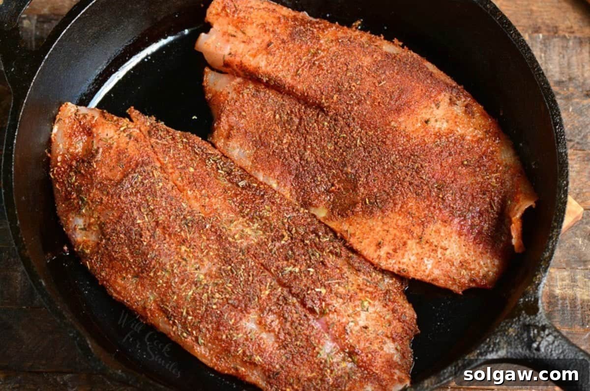 Two uncooked blackened tilapia fillets in a cast iron skillet, ready for cooking.