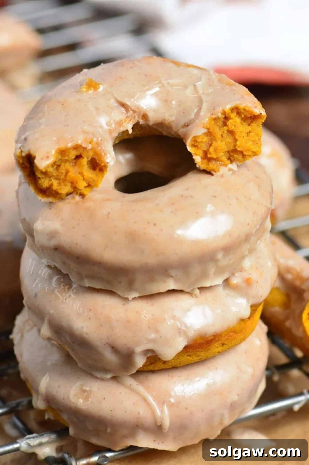 A close-up of a pumpkin donut torn in half, showcasing its soft interior, next to whole glazed donuts on a cooling rack.