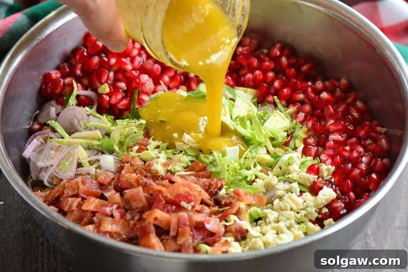 Mustard vinaigrette being poured from a glass jar into a large metal mixing bowl filled with the colorful Brussels sprouts salad ingredients, ready for tossing.