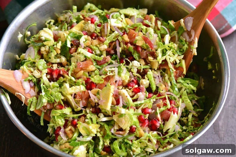 A large metal mixing bowl brimming with all the colorful ingredients of the Brussels sprouts salad, being gently tossed together to combine evenly before dressing.