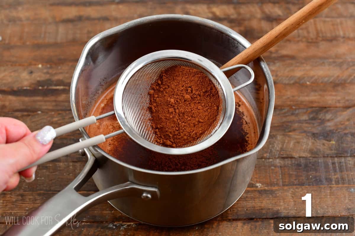 A fine mesh strainer sifting cocoa powder into a pot of warm milk on a stovetop.