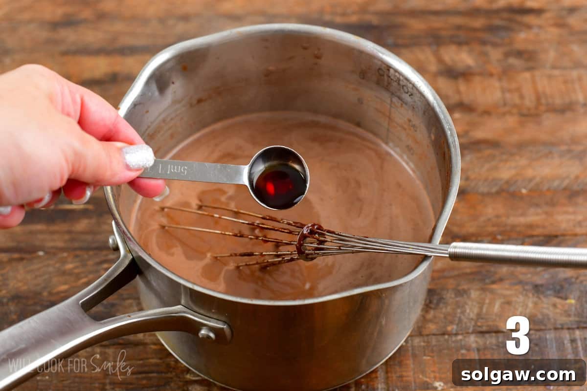 A measuring spoon adding vanilla extract to a pot of hot chocolate mixture on a wooden surface.