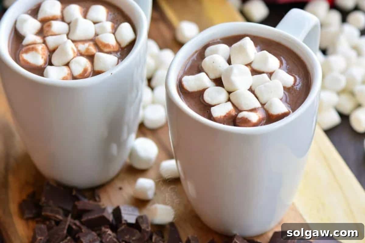 Two mugs of homemade hot chocolate, one topped with marshmallows, are presented on a wooden cutting board.