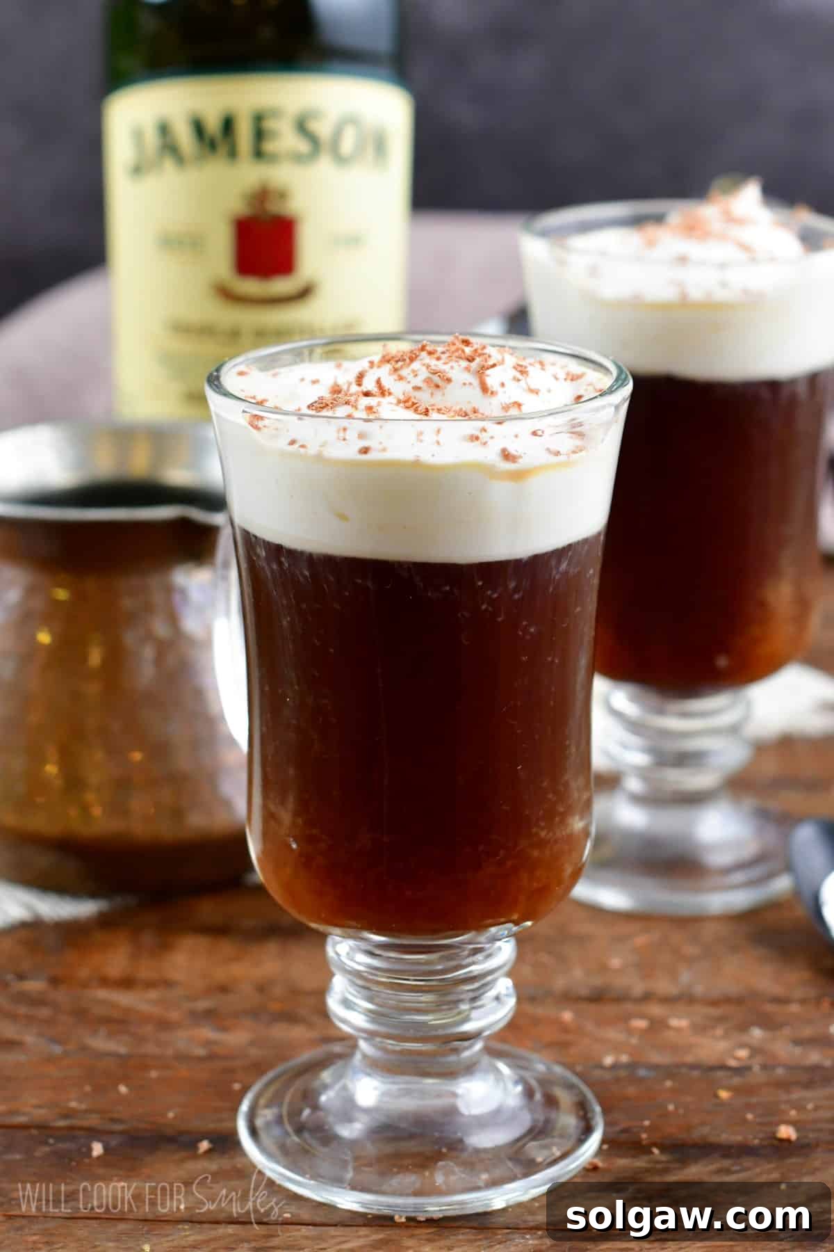 A glass filled with Irish coffee, topped with homemade whipped cream, with a bottle of Jameson and another glass blurred in the background.