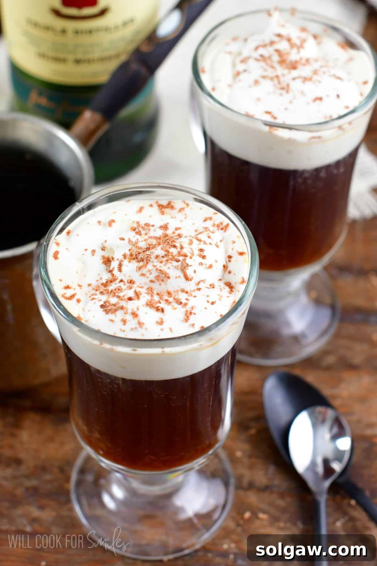Two mugs of Irish coffee, each crowned with luscious whipped cream, elegantly placed on a wooden surface with two spoons.