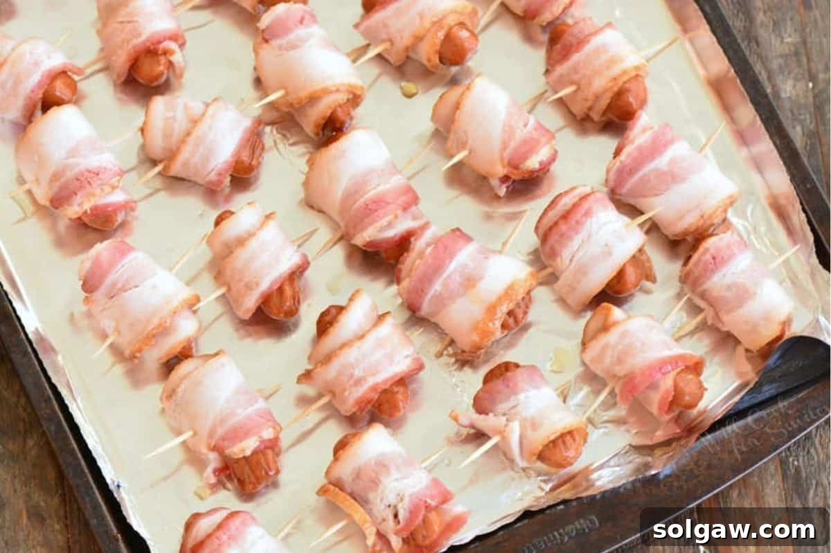 Several little sausages, each meticulously wrapped in bacon and secured with a toothpick, arranged on a baking sheet before cooking.