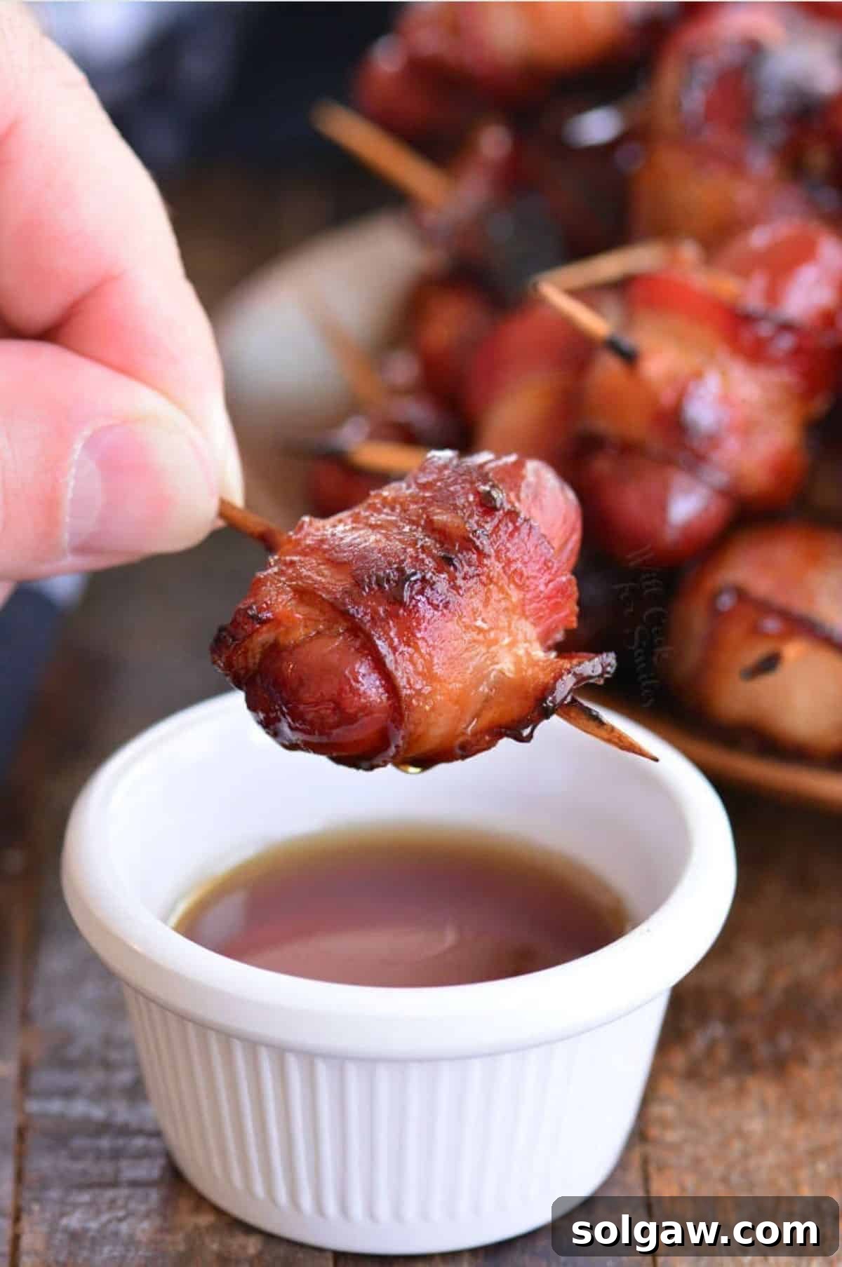A bacon-wrapped little smokie being dipped into a small bowl of golden maple syrup, showcasing the perfect sweet and savory combination.
