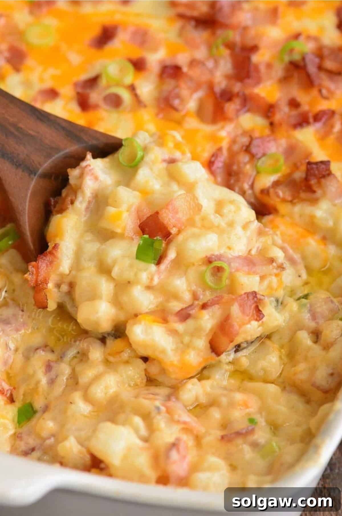 A bubbling, golden-brown baked loaded cheesy potato casserole in a large baking dish. A wooden spoon is gently scooping out a generous portion, revealing layers of creamy potatoes, melted cheese, and crispy bacon bits.
