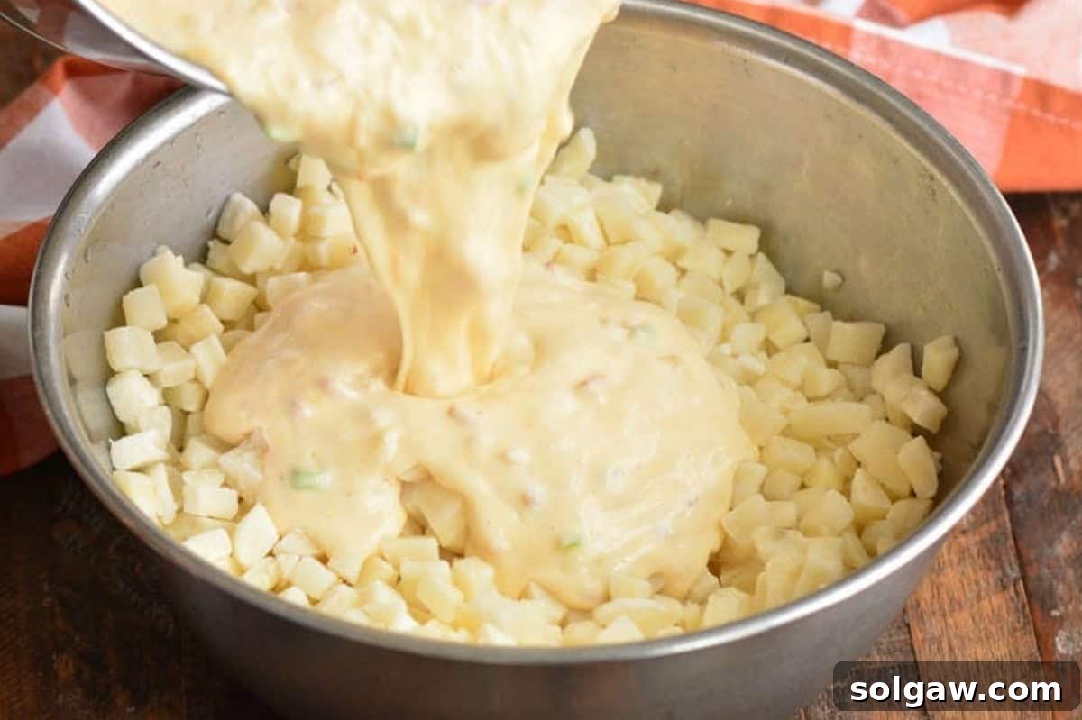 Pouring rich, golden cheese sauce over a bowl of diced hash brown potatoes, ready to be mixed for the ultimate loaded cheesy potato casserole.