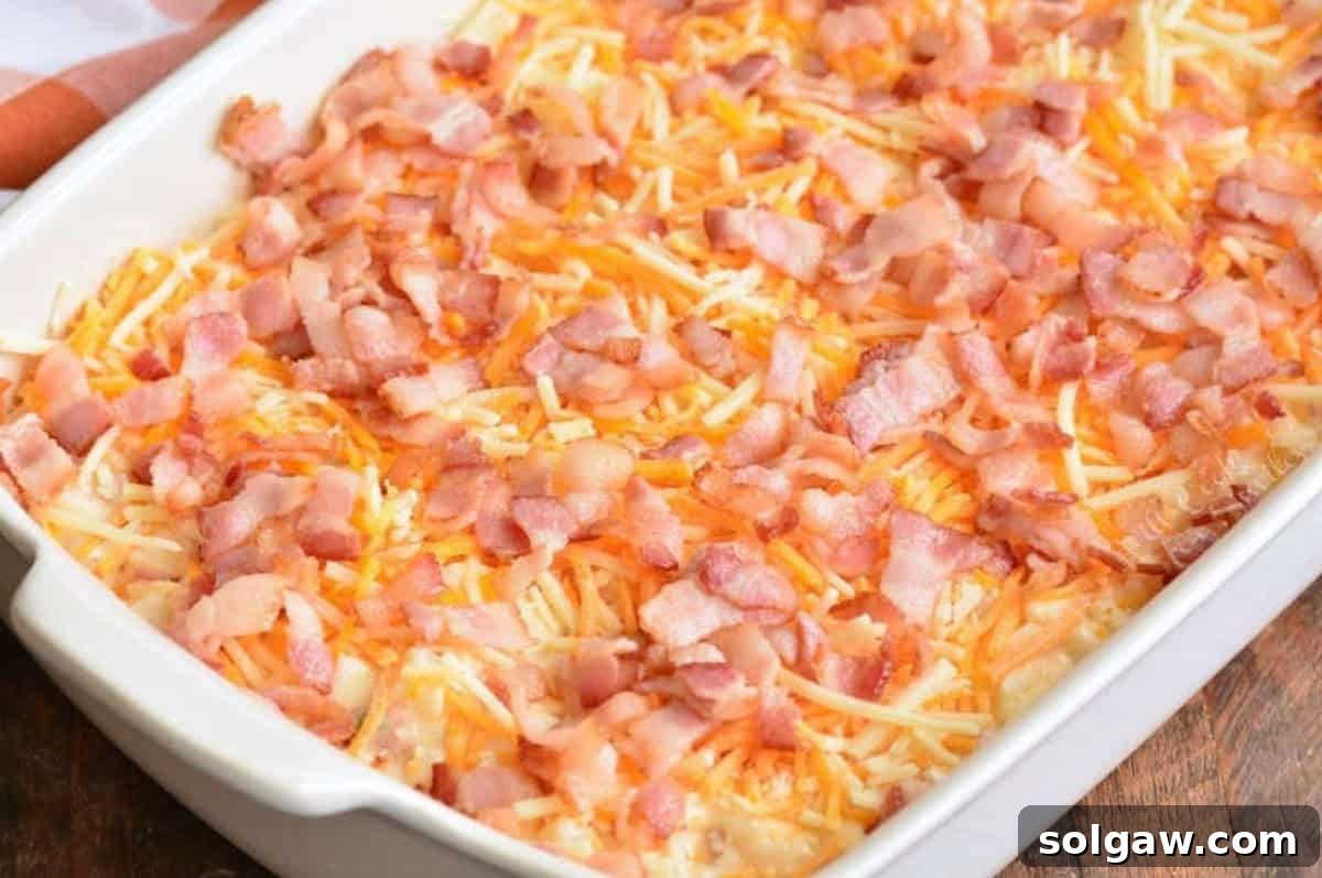 An unbaked loaded cheesy potato casserole, topped generously with crispy bacon bits and shredded cheddar cheese, ready to go into the oven.