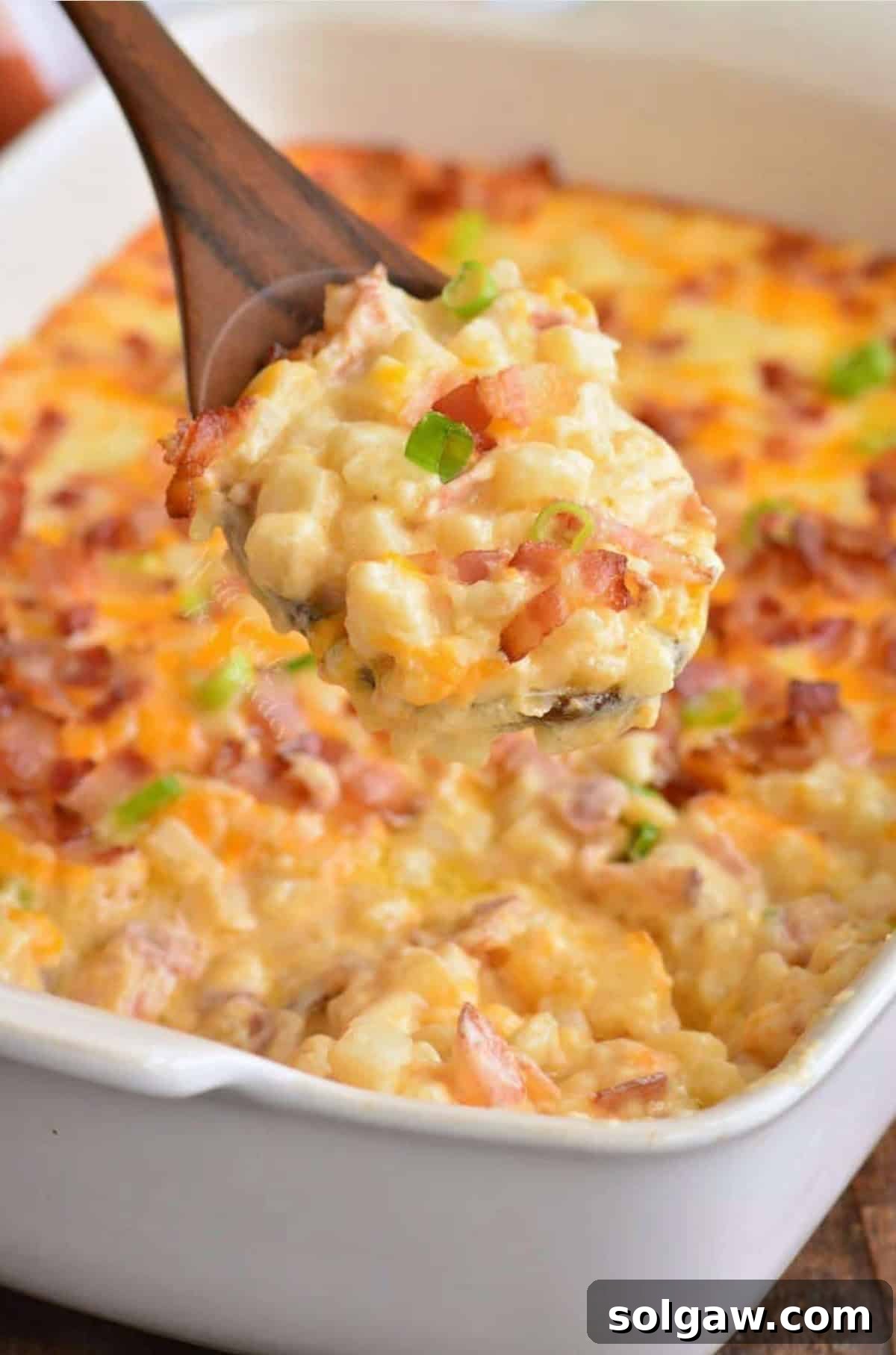 A close-up shot of creamy, cheesy loaded potato casserole being scooped onto a wooden spoon, showcasing the rich texture, melted cheese, and crispy bacon bits.