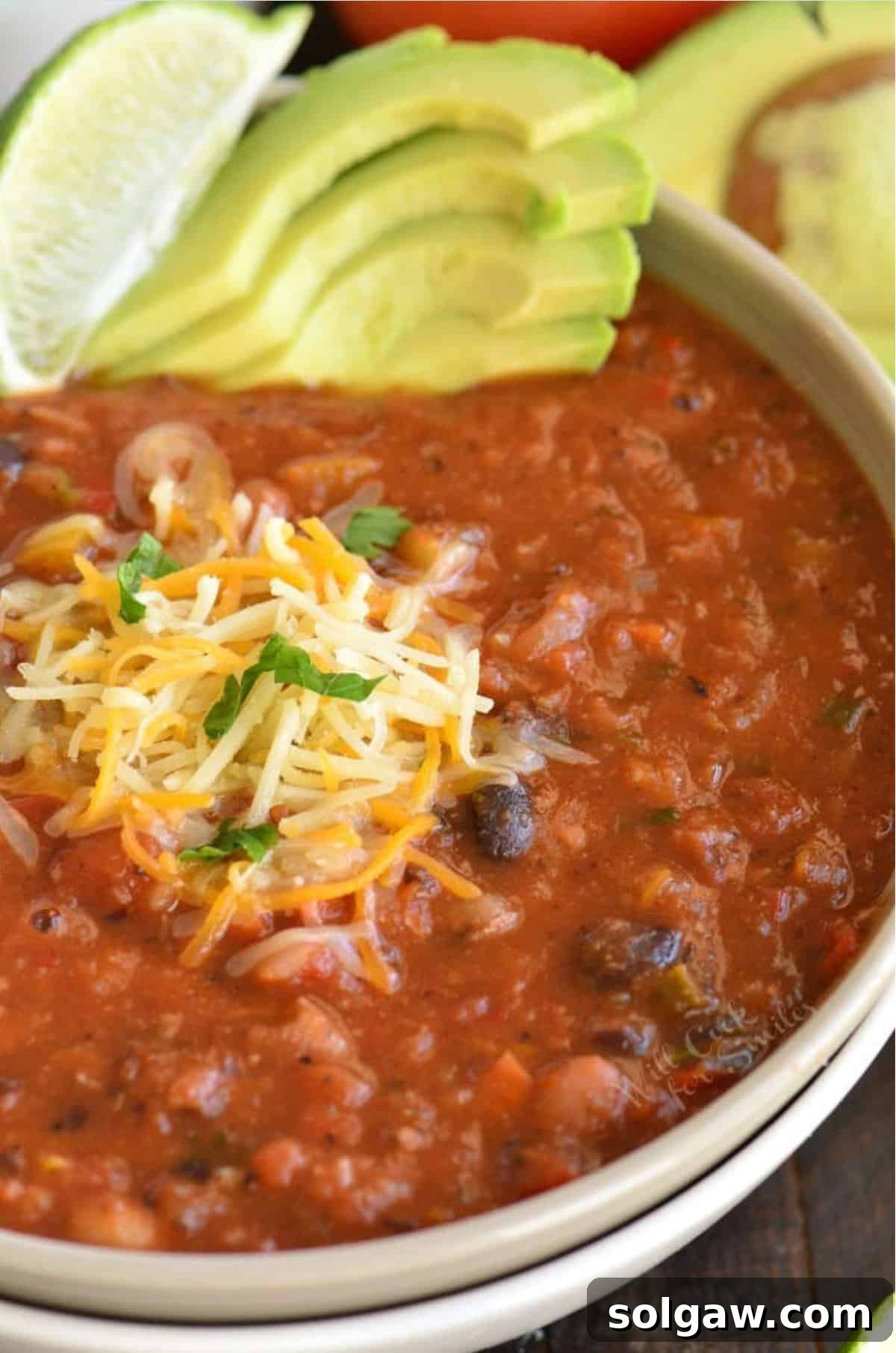A hearty bowl of vegetarian chili, garnished with shredded cheese, a fresh lime wedge, and sliced avocado.
