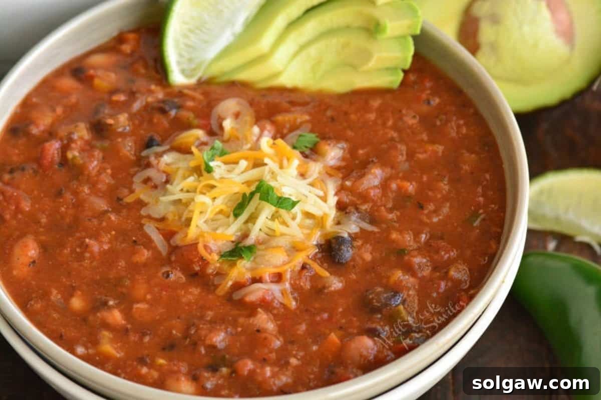 A bowl of vegetarian chili garnished with shredded cheese, a lime wedge, and sliced avocado, with a half avocado on the side.