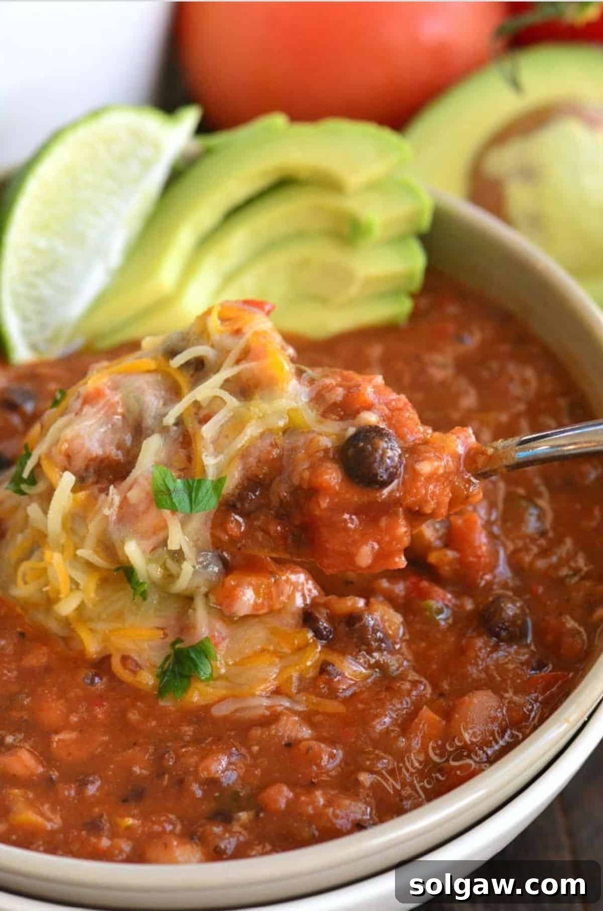 A bowl of vegetarian chili garnished with shredded cheese, a lime wedge, and sliced avocado, served with a spoon.