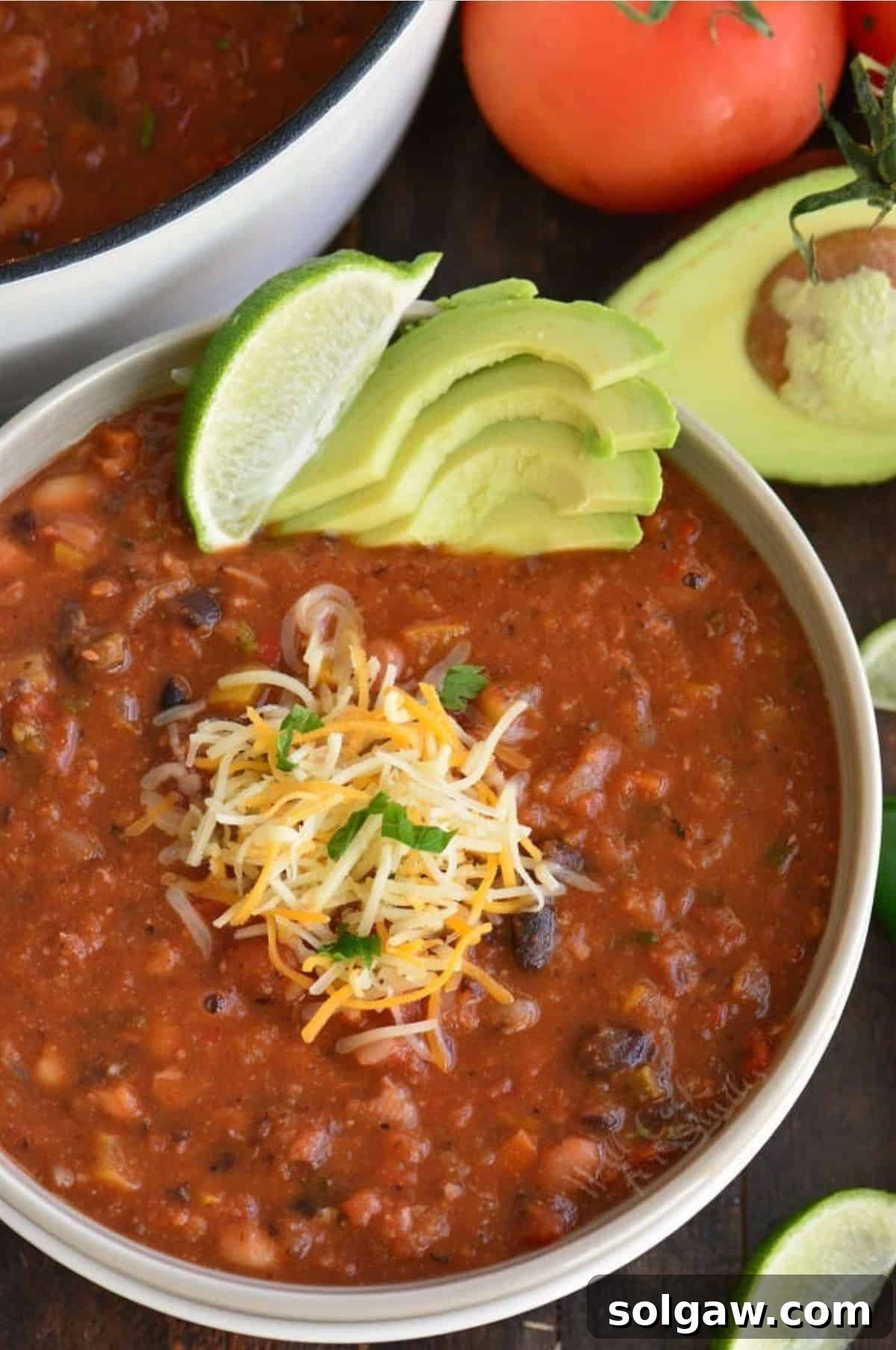 A bowl of vegetarian chili garnished with shredded cheese, a lime wedge, and sliced avocado, with fresh tomato and avocado in the background.