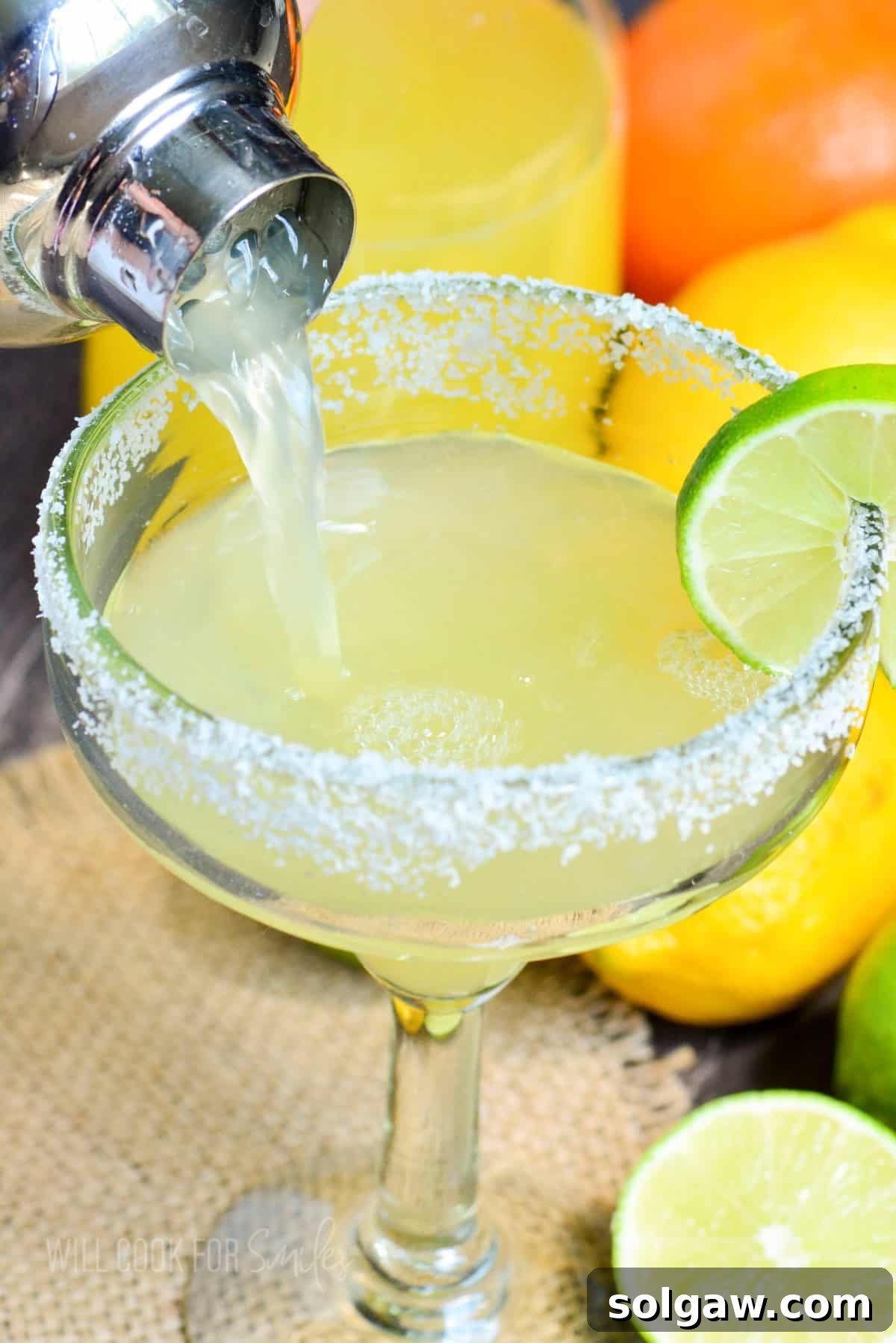 Close-up of a hand pouring a light-colored margarita cocktail into a chilled glass with a perfectly salted rim, ready for enjoyment.