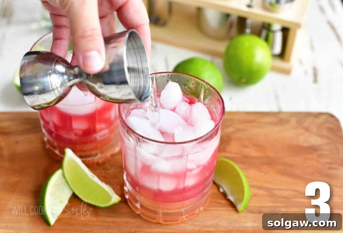 A hand pouring clear liquor from a jigger into a glass already containing pink and yellow juices, preparing a Bay Breeze cocktail.