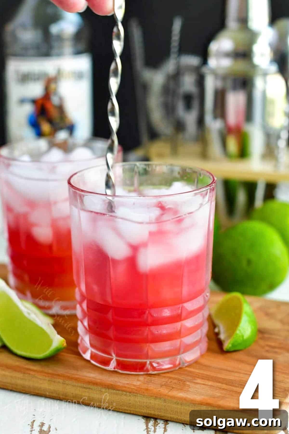 A Bay Breeze cocktail being stirred with a long bar spoon, showing its vibrant pink hue and ice, with lime wedges arranged around.