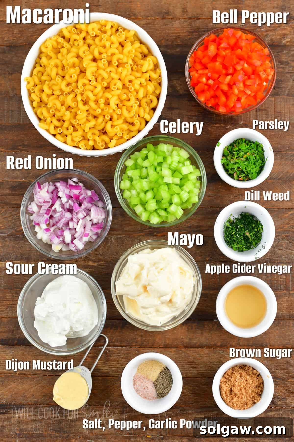 Homestyle Macaroni Salad 3 Fresh ingredients for a homemade macaroni salad, including elbow macaroni, red onion, celery, bell pepper, and fresh herbs, artfully arranged on a wooden board.