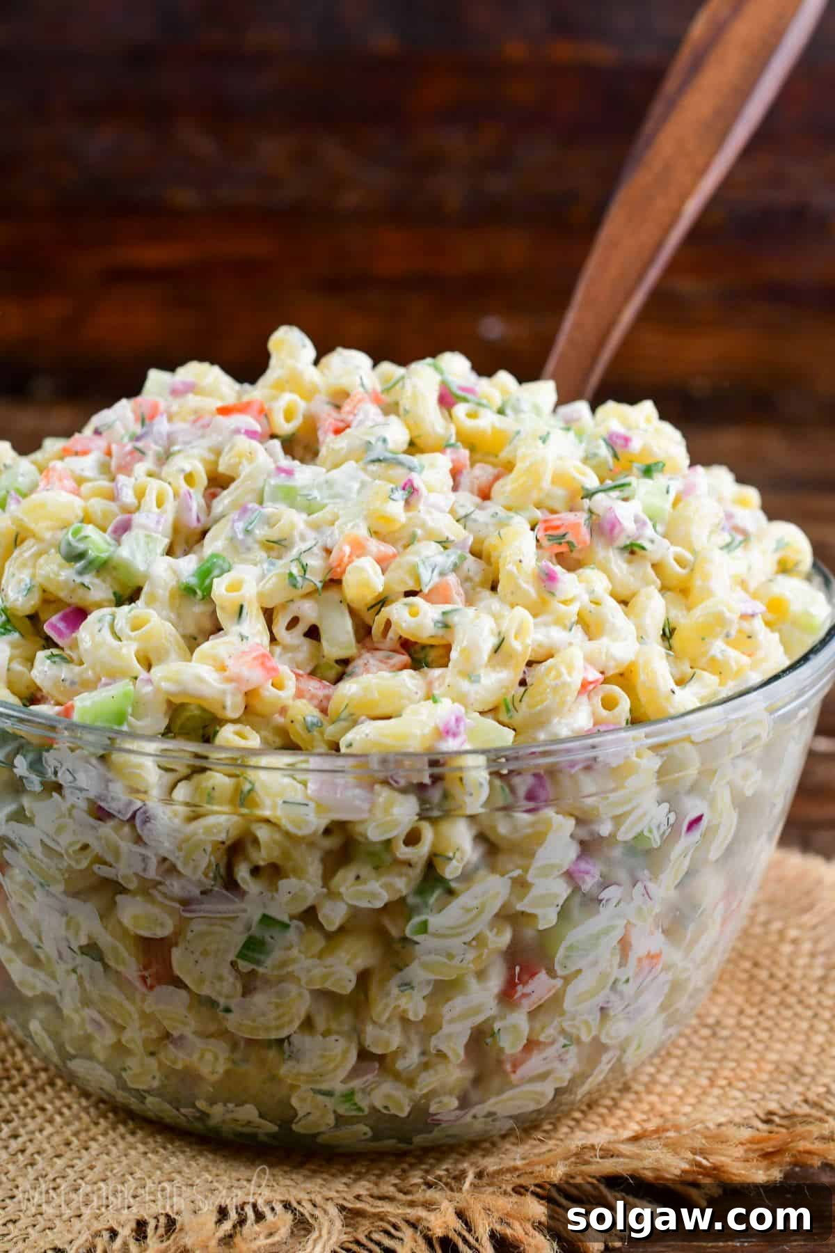 Homestyle Macaroni Salad 7 A large, inviting glass bowl filled with creamy macaroni salad, showcasing its vibrant mix of pasta and vegetables, served with a wooden spoon.