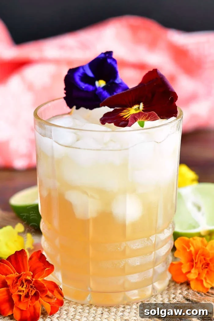 closeup of glass filled with crushed ice and light yellow cocktail decorated with purple and burgundy flowers