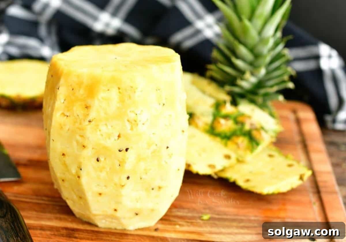 A skinned pineapple standing on a cutting board, with the removed top, bottom, and skin pieces behind it, showing the clean, edible fruit.