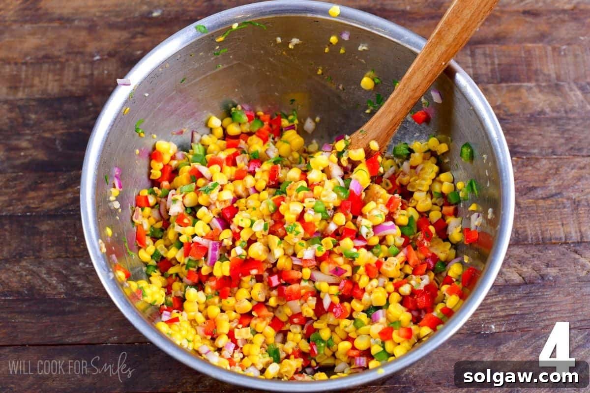A wooden spoon gently mixes the colorful corn salsa ingredients in a metal bowl, blending the flavors together.