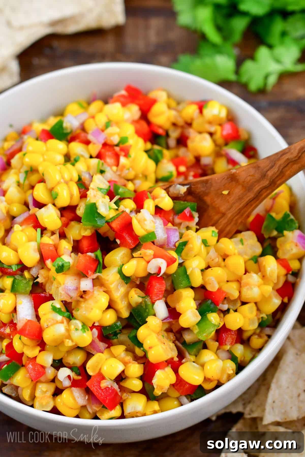 A generous scoop of vibrant corn salsa is lifted with a wooden spoon from a white serving bowl, showcasing its fresh appeal.