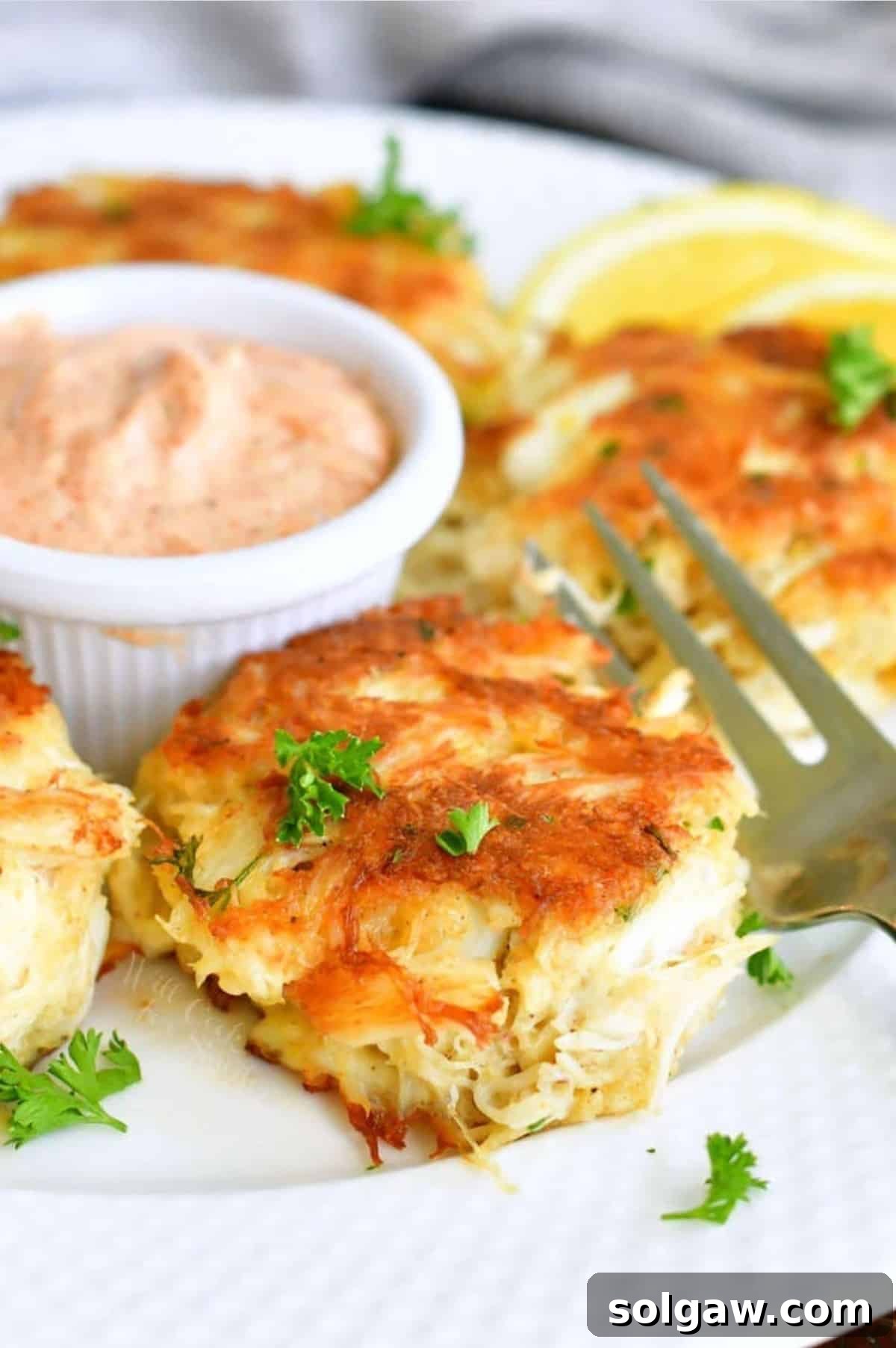 Delicately pan-fried crab cake on a white plate, served with a fork and creamy remoulade sauce in a small white cup, garnished with fresh herbs.