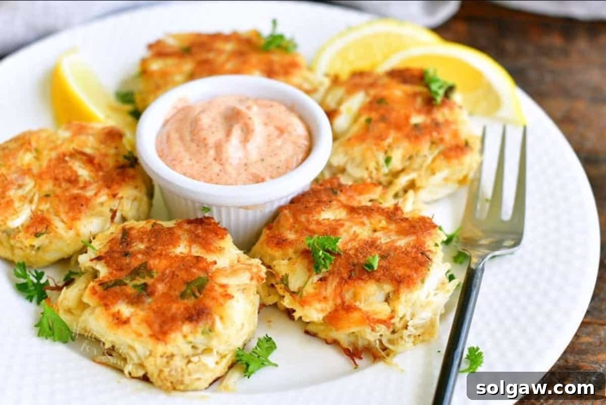 Five golden-brown crab cakes artfully arranged around a small bowl of pink remoulade sauce, with a fork and fresh lemon wedges, ready for serving.