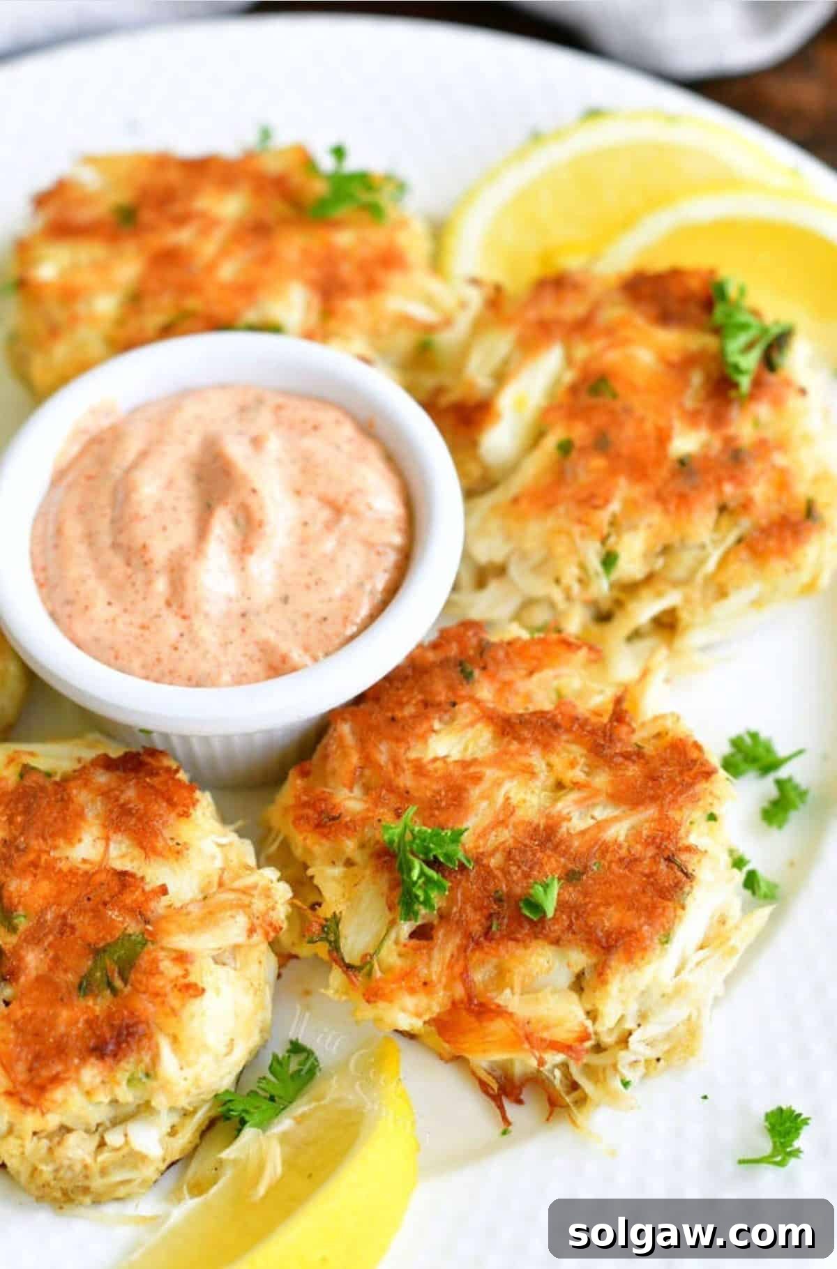 Four perfectly cooked crab cakes displayed on a pristine white plate, accompanied by a side of creamy remoulade sauce and vibrant lemon wedges, ready to be enjoyed.