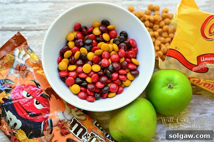 M&M bag and m&m's in a white bowl with 2 whole green apples to the bottom of it and a bag of caramel chips with some spilled out on the table 