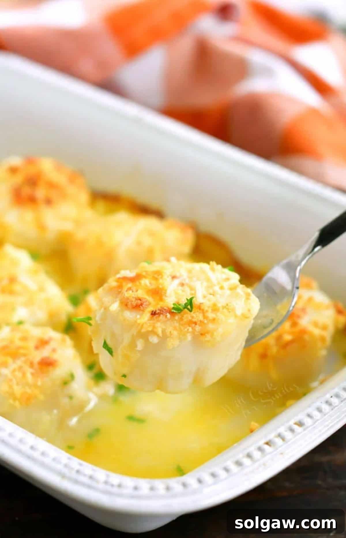 A single perfectly baked scallop held on a fork, glistening over a baking dish filled with more scallops.