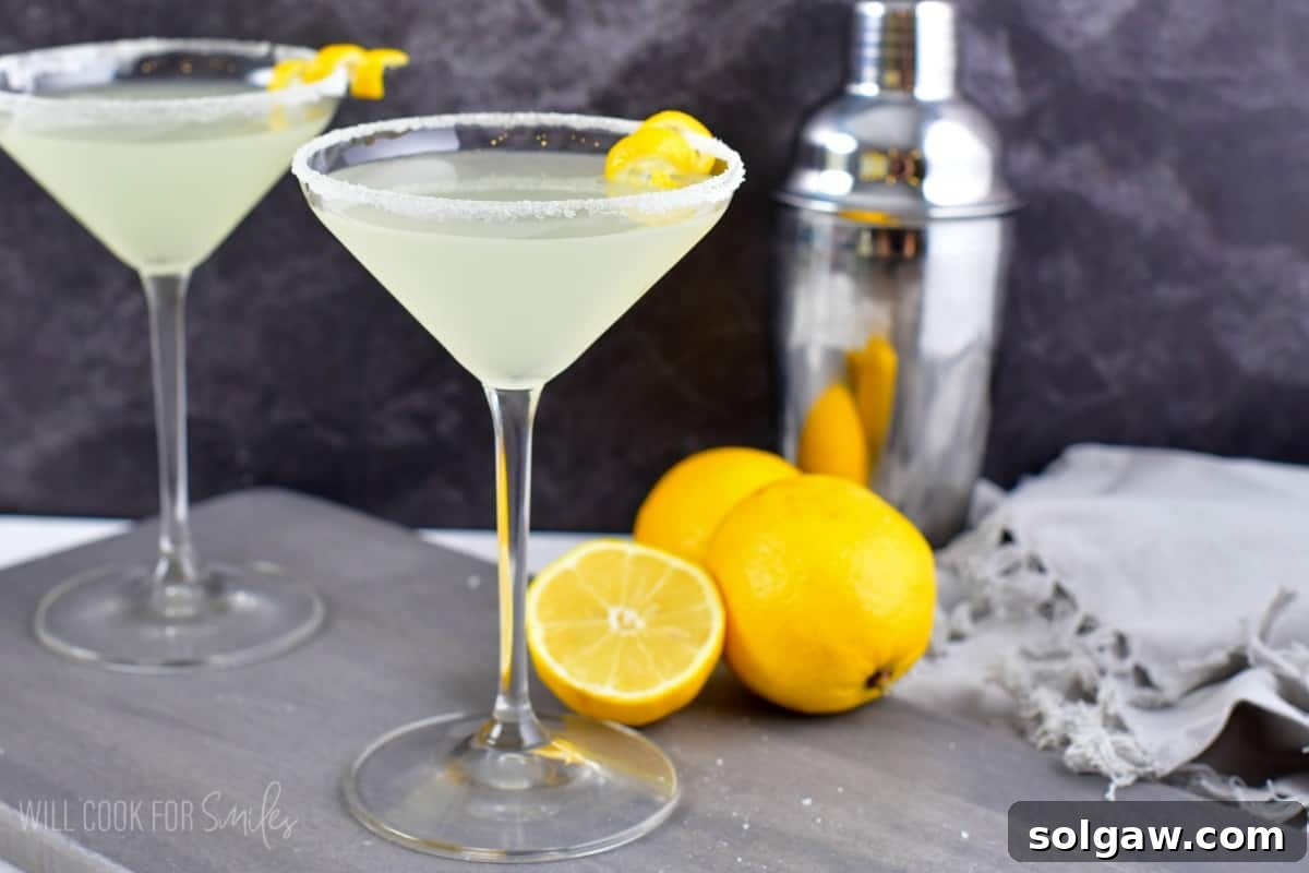 Lemon drop in a martini glass with a sugar rimmed glass and two whole lemons and one half of a lemon on a wood surface.