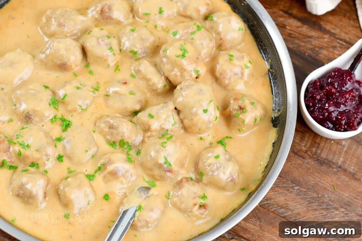 Creamy Swedish Meatballs 9 A pan full of rich Swedish meatballs in creamy gravy sits invitingly next to a small, vibrant serving dish of lingonberry jam, showcasing a complete meal.