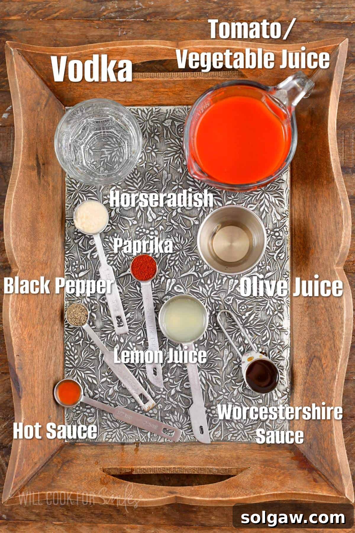 Labeled ingredients for Bloody Mary on a wooden and silver tray, including V8, vodka, horseradish, spices, and garnishes.