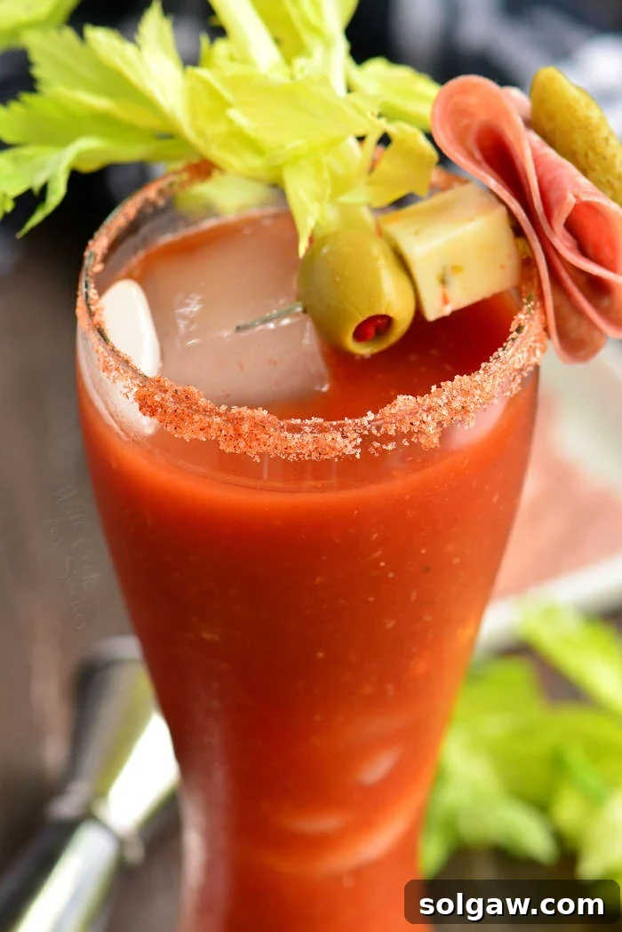Close-up of a V8 Bloody Mary cocktail in a tall glass with a salted rim and a skewer of garnishes including an olive, cheese cube, folded salami, and a pickle.