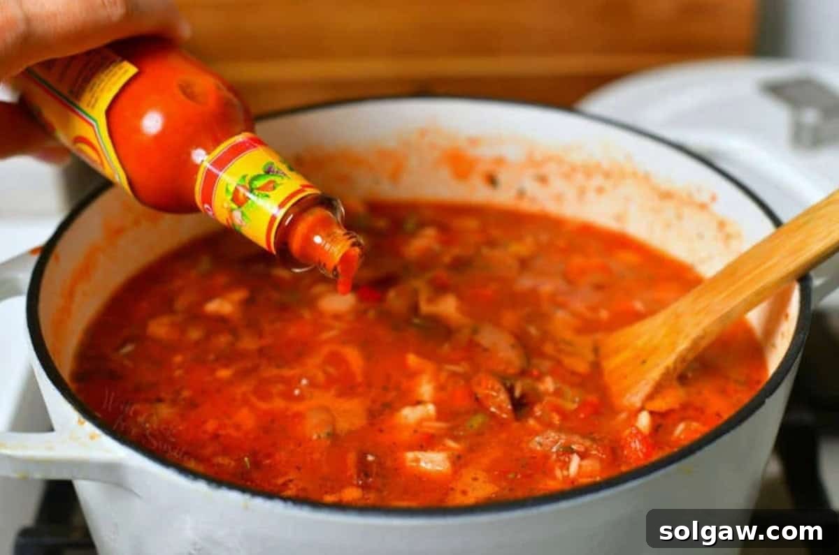 A few drops of hot sauce being added to a pot of Jambalaya simmering on the stove, enhancing its authentic spicy flavor.
