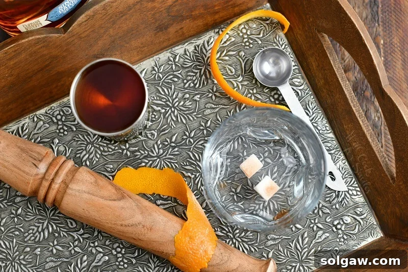 overhead photo of cocktail muddler, jigger of bourbon, sugar cubes, orange peel