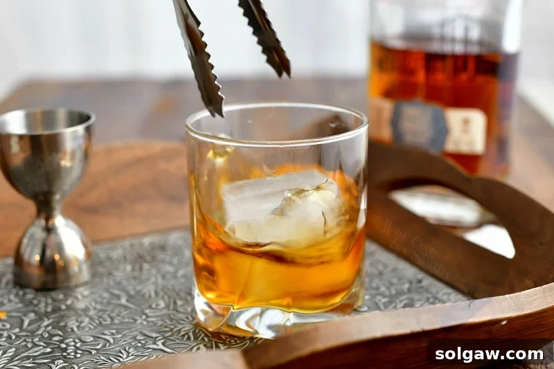 side view: metal tongs dropping ice cube into whiskey cocktail