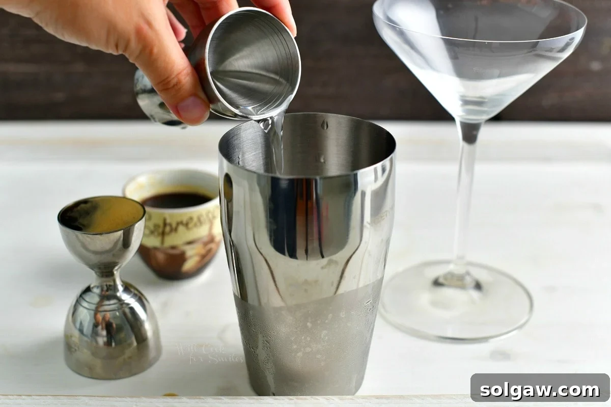 A hand pouring clear vodka from a bottle into a cocktail shaker filled with ice.