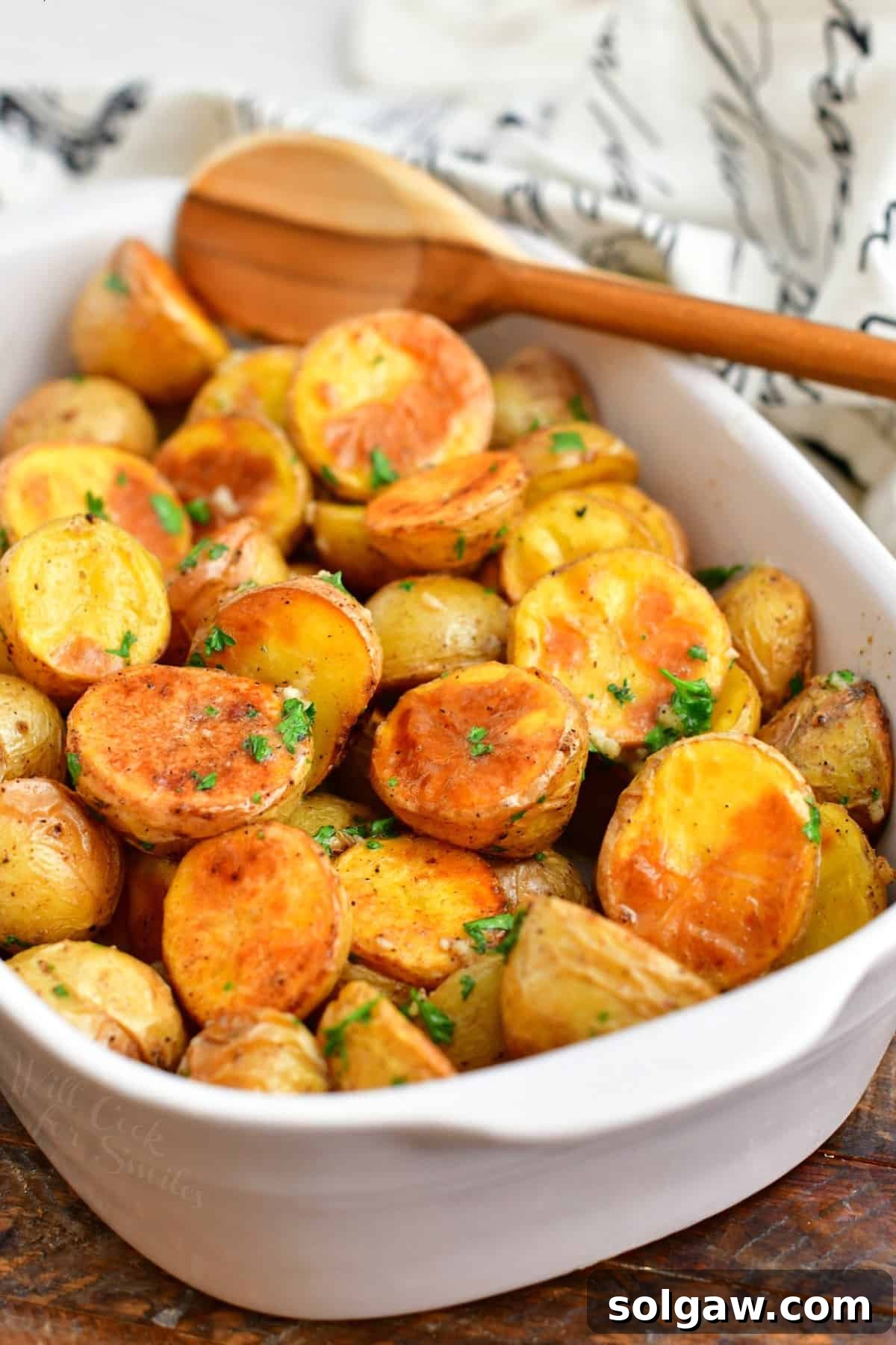 A wooden spoon resting in a white casserole dish filled with generously coated roasted potatoes, ready to be served.