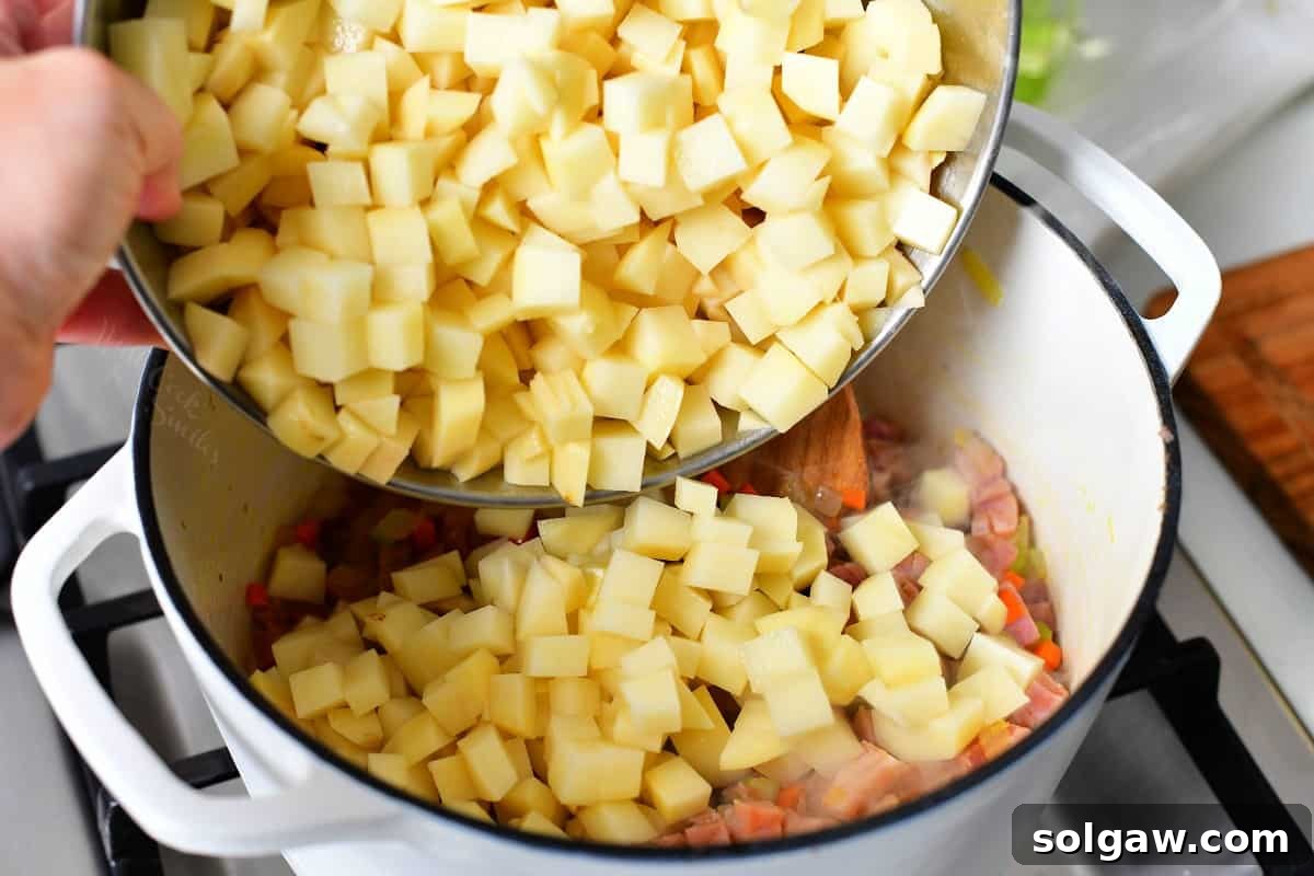 Diced potatoes being added to a pot already containing sautéed vegetables and ham for soup.